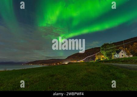 Splendida aurora boreale verde sopra un fiordo e una casa sull'isola di Kvaloya vicino a Tromsø. luci polari danzanti su una montagna, aurora boreale Foto Stock