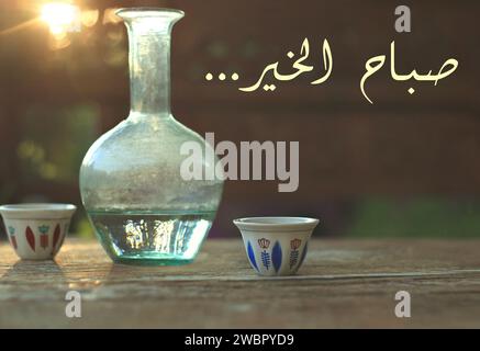 Buon giorno scritto in arabo. Due tazze di caffè tradizionali libanesi insieme a una caraffa d'acqua in vetro, ebrik. Foto Stock