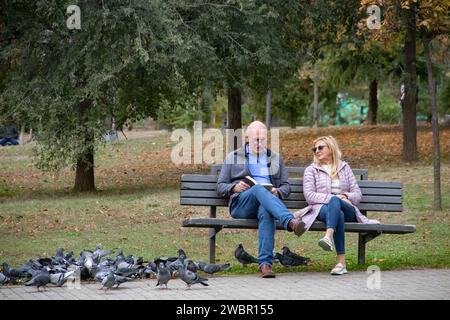 Colpo di stato anziano di mezza età [le godendo una bella giornata di sole autunnale alla panchina nel parco pubblico della città, con un gregge di piccioni intorno a loro Foto Stock