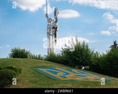 Monumento della madrepatria Ucraina contro un cielo con nuvole e l'emblema di stato dell'Ucraina in primo piano Foto Stock