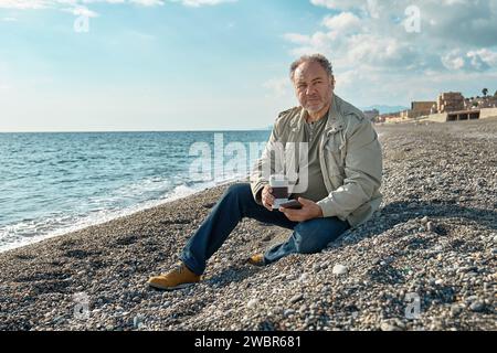 Uomo con la barba di mezza età che usa lo smartphone mentre è seduto al mare in inverno o in primavera. Foto Stock