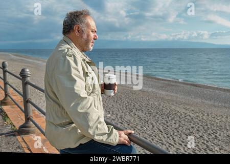 Uomo penoso di mezza età barbato che beve caffè al mare in inverno o in primavera. Foto Stock