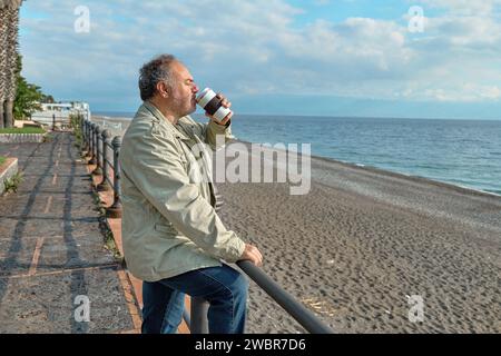 Uomo penoso di mezza età barbato che beve caffè al mare in inverno o in primavera. Foto Stock