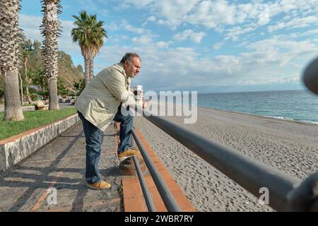 Uomo penoso di mezza età barbato che beve caffè al mare in inverno o in primavera. Foto Stock