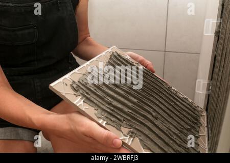 Donna che affianca le pareti del bagno, applica l'adesivo su una piastrella e posiziona la piastrella in ceramica nella giusta posizione. Processo di installazione delle piastrelle nel bagno Foto Stock