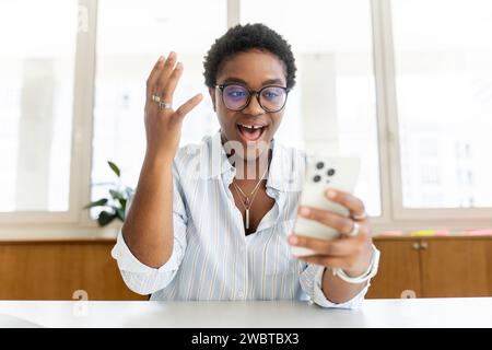 Felicissima donna afro-americana che guarda lo schermo del telefono, celebra il successo. Eccitata soddisfatta giovane dipendente in ufficio con smartphone, leggendo buone notizie in messaggi, vincita alla lotteria, offerta di lavoro Foto Stock