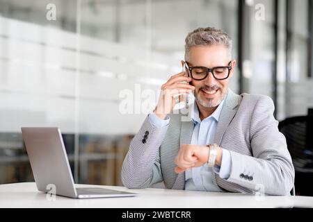 Uomo d'affari ottimista senior degli anni '60 che chiama sullo smartphone un orologio da polso o un orologio intelligente in mano seduto sul posto di lavoro in un ufficio moderno. Puntualità e concetto di persone Foto Stock