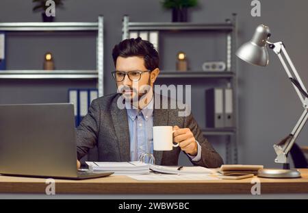 Un giovane serio e concentrato con occhiali, vestito in stile ufficio, è seduto alla sua scrivania in ufficio a bere tè o caffè e a scrivere testo sul computer portatile. Foto Stock