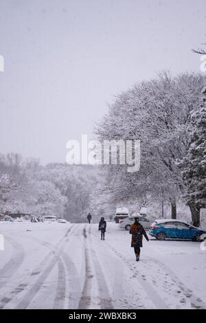 Un gruppo di individui può essere visto passeggiare attraverso un paesaggio innevato, con una fila di veicoli visibili sullo sfondo Foto Stock