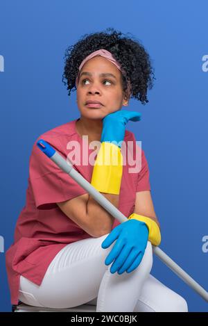 Donna di pulizia afro latina stanca anni '40 che indossa camicia magenta e guanti in gomma gialli per la protezione delle mani, isolata su sfondo blu Foto Stock