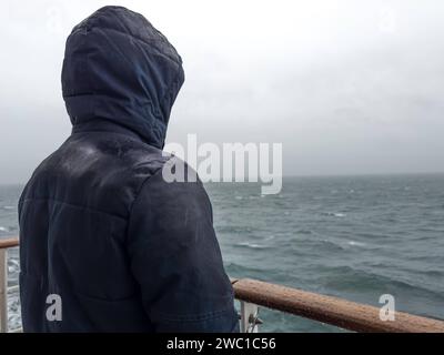 9 dicembre 2023: L'uomo sta con la giacca sulla ringhiera di una nave da crociera e guarda il mare sotto la pioggia **** Mann steht mit Jacke an der Rehling von einem Kreuzfahrtschiff und schaut bei Regen auf das Meer hinaus Foto Stock