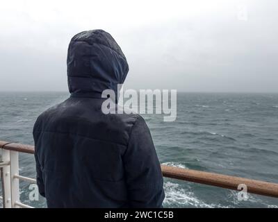 9 dicembre 2023: L'uomo sta con la giacca sulla ringhiera di una nave da crociera e guarda il mare sotto la pioggia **** Mann steht mit Jacke an der Rehling von einem Kreuzfahrtschiff und schaut bei Regen auf das Meer hinaus Foto Stock