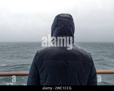 9 dicembre 2023: L'uomo sta con la giacca sulla ringhiera di una nave da crociera e guarda il mare sotto la pioggia **** Mann steht mit Jacke an der Rehling von einem Kreuzfahrtschiff und schaut bei Regen auf das Meer hinaus Foto Stock