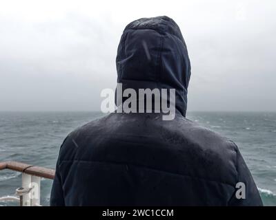 9 dicembre 2023: L'uomo sta con la giacca sulla ringhiera di una nave da crociera e guarda il mare sotto la pioggia **** Mann steht mit Jacke an der Rehling von einem Kreuzfahrtschiff und schaut bei Regen auf das Meer hinaus Foto Stock