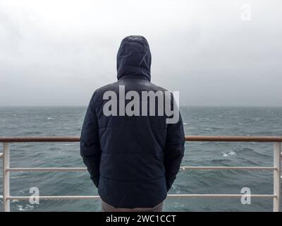 9 dicembre 2023: L'uomo sta con la giacca sulla ringhiera di una nave da crociera e guarda il mare sotto la pioggia **** Mann steht mit Jacke an der Rehling von einem Kreuzfahrtschiff und schaut bei Regen auf das Meer hinaus Foto Stock