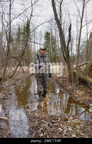 Un cacciatore si fa strada attraverso una pozzanghera nella foresta primaverile. Ha un fucile a pistola sulla spalla. Il crepuscolo serale si approfondisce. Foto Stock