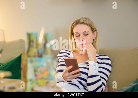 Attraente donna bionda di mezza età seduta sul divano e che usa lo smartphone Foto Stock