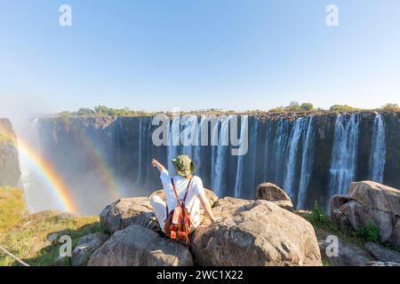 Donna seduta su una roccia punta a Victoria Falls - Zimbabwe, concetto di viaggio Foto Stock