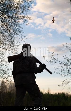 Un cacciatore con un fucile scaricato sulla spalla si trova al crepuscolo in una radura forestale. Guarda il becco maschio, che fa un corteggiamento Foto Stock