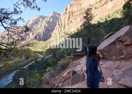 Una turista esplora un sentiero nel parco nazionale di zion con la sua fotocamera del telefono. Scatta una foto del fiume vergine e delle vicine vette montane. Foto Stock