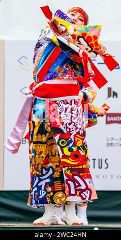 Primo piano, ballerina giapponese di yosakoi che usa naruko, clappers, in luminoso costume multicolore sul palco all'aperto, festival Kumamoto Kyusyu Gassai. Foto Stock