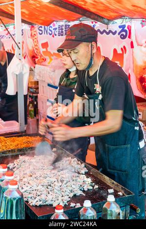 Chiosco di fast food in Giappone durante il festival primaverile della fioritura dei ciliegi. Uomo che usa due spatole di metallo per preparare il cibo su una piastra calda accanto a un vassoio di nozze. Foto Stock