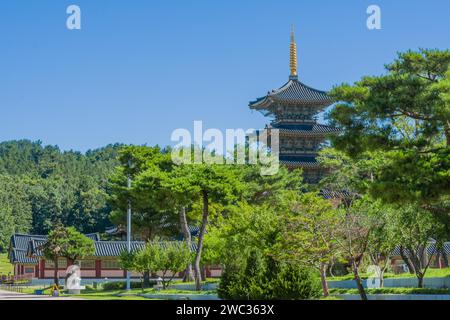 Alta pagoda orientale con tetto piastrellato e guglia dorata e vista parziale degli edifici circostanti dietro splendidi alberi nel parco pubblico nelle giornate di sole Foto Stock