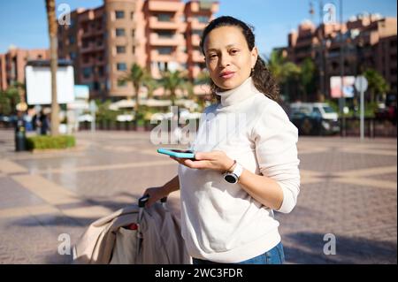 Giovane bella donna viaggiatrice con valigia, in abiti informali utilizzando smartphone all'aperto, controllando le indicazioni sulla mappa online, cercando con sicurezza un Foto Stock