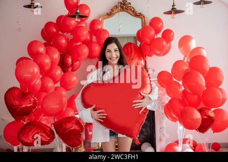 Giovane donna sorridente in camera decorata per festeggiare San Valentino. La ragazza tiene un grande palloncino rosso a forma di cuore. Petali di rosa sparsi Foto Stock