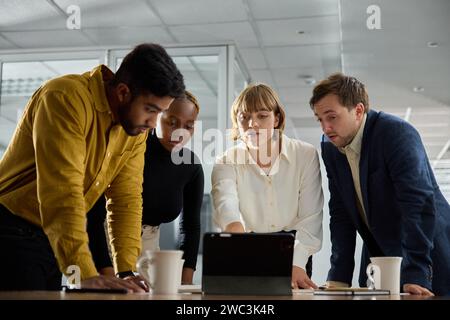 Quattro giovani adulti multirazziali che indossano abbigliamento da lavoro si incontrano intorno al tablet digitale in ufficio Foto Stock