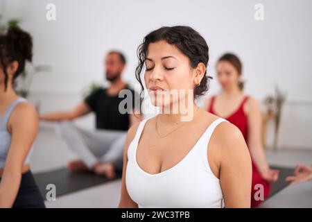 Primo piano giovane latina con gli occhi chiusi che pratica esercizi di yoga al chiuso. Gruppo di persone Foto Stock