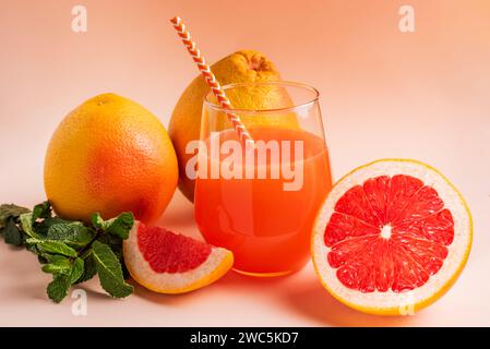 Diversi pompelmi con uno aperto a metà che mostra il suo interno accanto a un bicchiere con una cannuccia con il suo succo e la menta su sfondo rosa. Foto Stock