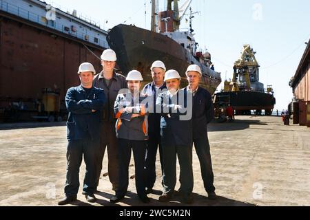 Il personale addetto alla manutenzione si pone di fronte alla grande nave presso il bacino di carenaggio del cantiere. Gli ingegneri con elmetti, le tute sono pronte per le riparazioni marittime. Industriale Foto Stock