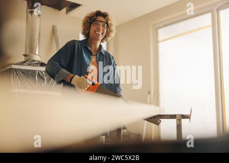 Una donna matura felice che indossa occhiali di sicurezza sta ristrutturando abilmente l'interno di una casa. Usando una sega a croce, sta tagliando legno per una casa fai da te i Foto Stock