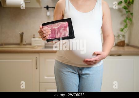 Una donna incinta tiene la carne di pancetta in mano mentre si trova in una cucina di casa. Gravidanza e corretta alimentazione in attesa di un bambino Foto Stock