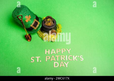 Vista dall'alto su uno sfondo verde e isolato si trovano l'iscrizione Happy Patrick's Day, Hat, leprechaun pot con monete d'oro, horseshoe per buona fortuna, pala Foto Stock