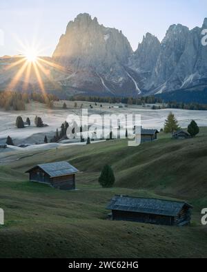 Foto verticale del sole che sorge sopra l'alto alpeggio con cottage da dietro un muro di montagne Foto Stock