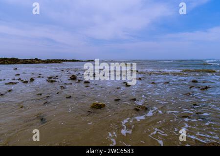Paesaggio di costa oceanica con onde che si innalzano sulle rocce nella sabbia e un piccolo affioramento roccioso sullo sfondo Foto Stock