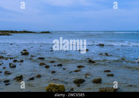 Paesaggio di costa oceanica con onde che si innalzano sulle rocce nella sabbia e un piccolo affioramento roccioso sullo sfondo Foto Stock