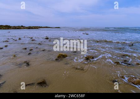 Paesaggio di costa oceanica con onde che si innalzano sulle rocce nella sabbia e un piccolo affioramento roccioso sullo sfondo Foto Stock