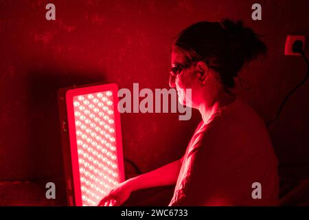 Donna anziana che fa la sessione di terapia a luce rossa con pannello e indossa una protezione per gli occhi a casa sul letto Foto Stock