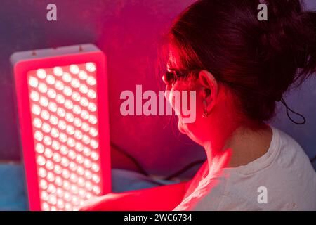 Donna anziana che fa la sessione di terapia a luce rossa con pannello e indossa una protezione per gli occhi a casa sul letto Foto Stock