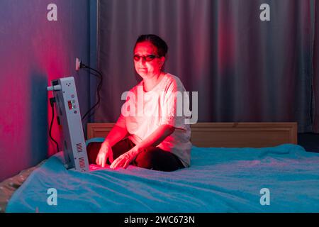 Donna anziana che fa la sessione di terapia a luce rossa con pannello e indossa una protezione per gli occhi a casa sul letto Foto Stock