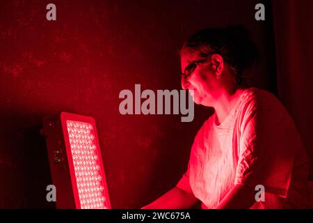 Donna anziana che fa la sessione di terapia a luce rossa con pannello e indossa una protezione per gli occhi a casa sul letto Foto Stock