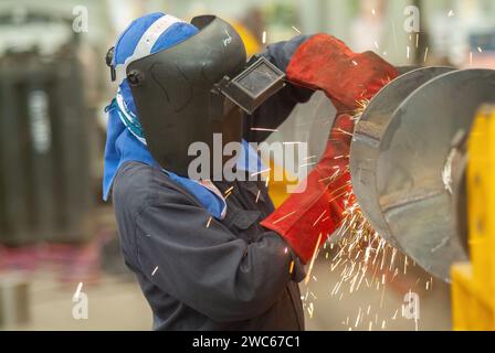 Lavoratore industriale che utilizza una smerigliatrice angolare nelle prime fasi di realizzazione di un trasportatore a vite in acciaio al carbonio. Foto Stock