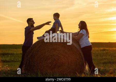 Una famiglia felice che gioca su un rotolo di paglia nella luce dorata della sera. Foto Stock