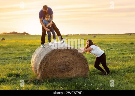 Divertiti con giochi per tutta la famiglia su un prato rurale all'aperto la sera al tramonto. Una madre spinge un rotolo di fieno e vuole che suo marito e suo figlio cadano. Un fami armonioso Foto Stock