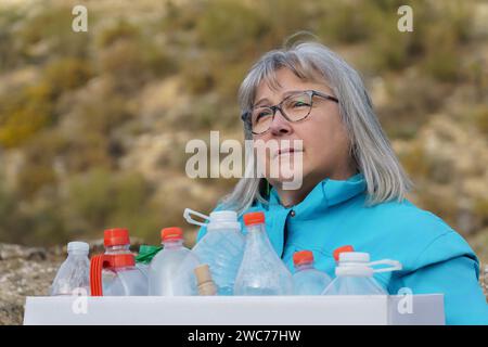 donna anziana dai capelli bianchi che tiene una scatola di cartone piena di bottiglie di plastica vuote raccolte sul campo per il riciclaggio, concetto di ecologia Foto Stock