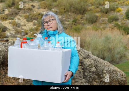 donna anziana dai capelli bianchi che tiene una scatola di cartone piena di bottiglie di plastica vuote raccolte sul campo per il riciclaggio, concetto di ecologia Foto Stock