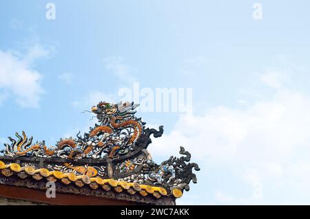 Una tradizionale scultura di drago, dettagliatamente dettagliata, si trova in cima a un'antica casa vietnamita, a simboleggiare potere e prosperità sotto un blu chiaro Foto Stock